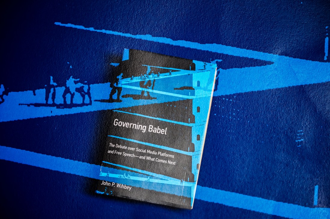A book titled 'Governing Babel: The Politics of Social Media Platforms and Free Speech' by John P. Wilson, displayed on a blue architectural surface with geometric shadows cast by structural beams.