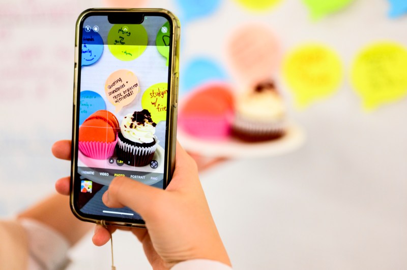 A person takes a photo of cupcakes decorated with colorful frosting and speech-bubble notes about happiness.