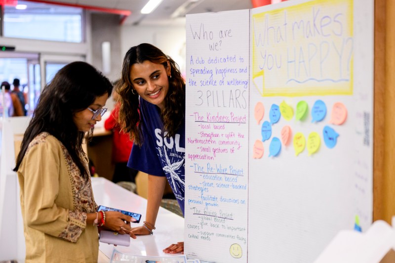 Two students smile while talking at a table beside posters about spreading happiness at the Happiness Cup Club event.