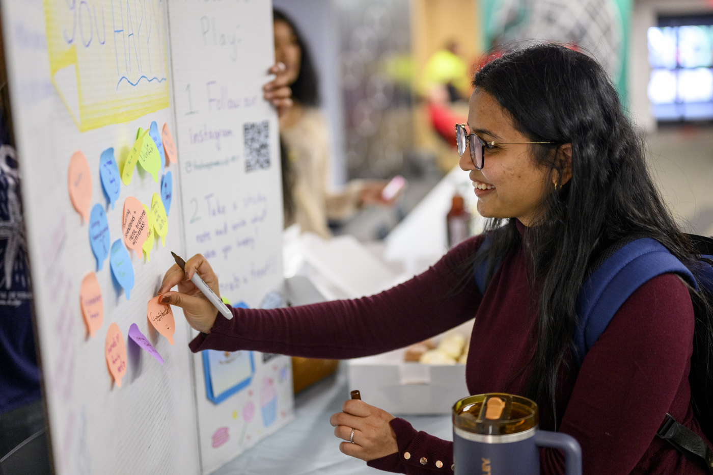 A student sticks their sticky note to a board asking people to write what makes them happy. 