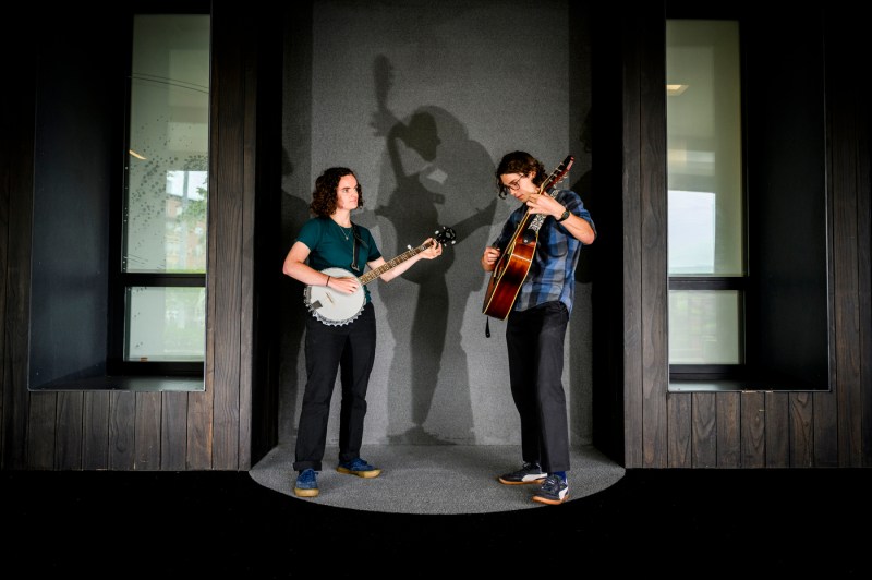 Two people stand on a stage. One plays the banjo while another plays a guitar.