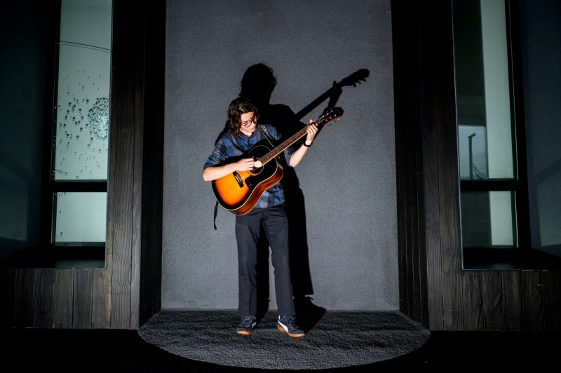 Garrett Compston standing in front of a grey background playing the guitar. 