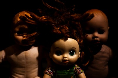 Three vintage dolls positioned in a triangle formation in dim lighting against a dark background, creating an unsettling atmosphere.