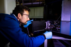 A man with glasses dressed in a blue lab coat tinkers with an electric panel in a lab.