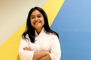 A headshot of Anjali Laddha, shown smiling in a white shirt against a blue and yellow background.