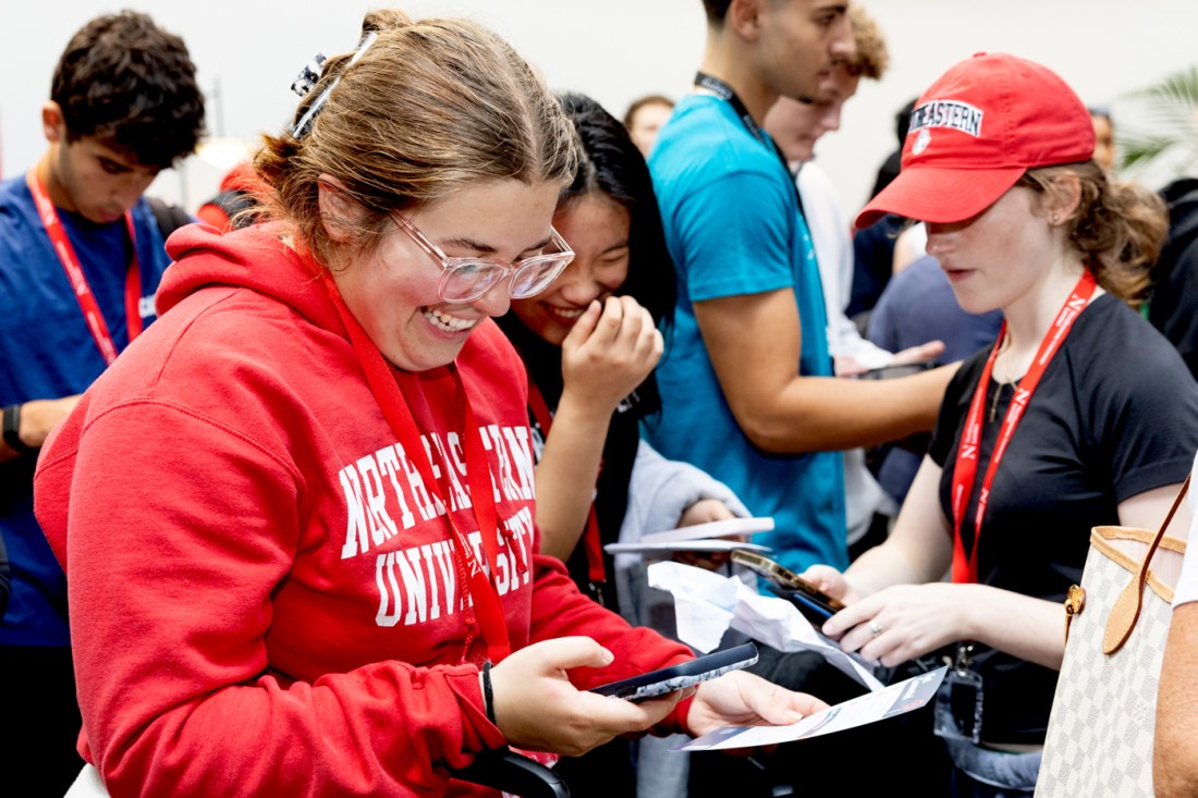 An excited student wearing a red 'Northeastern University' hoodie holds a pamphlet in one hand and their phone in the other.