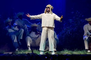 A photo of Bad Bunny, dressed in all white and sunglasses, performing on stage under a dark blue light.