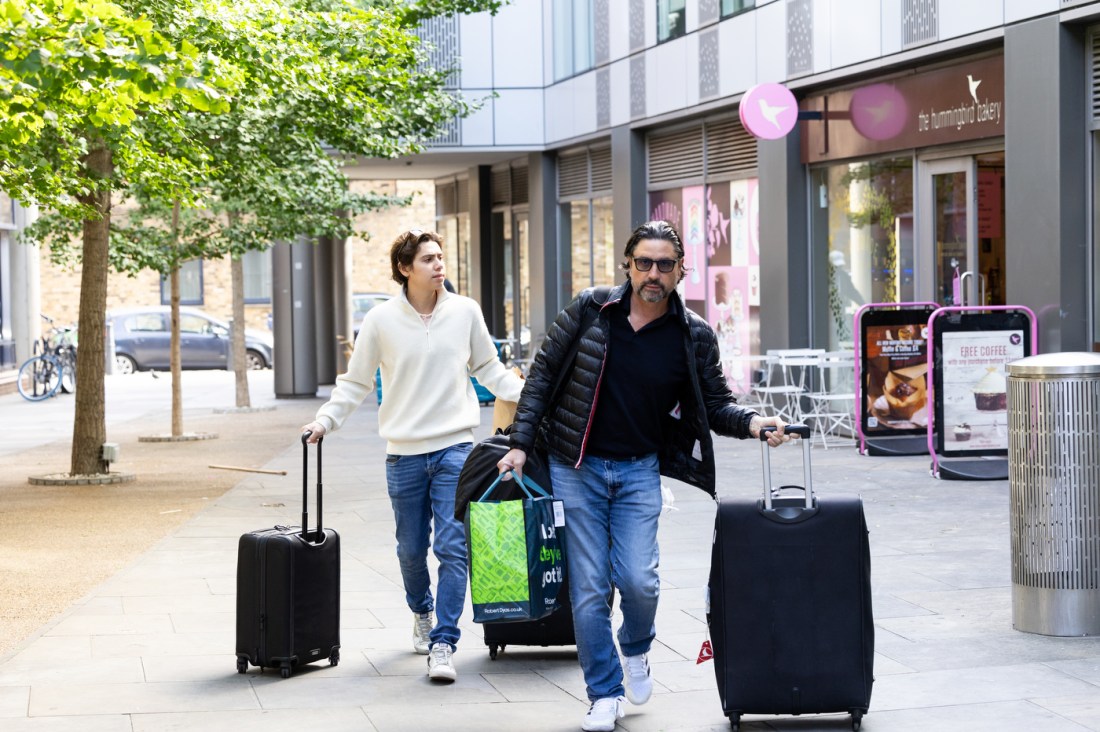 Two people wheeling large suitcases outside of a dorm building in London.