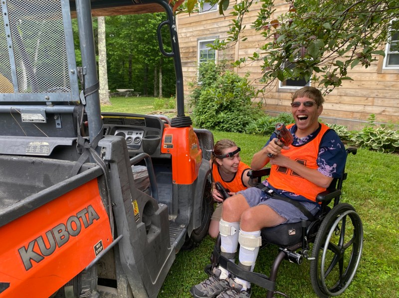 A person wearing an orange vest sits in a wheel chair next to a utility vehicle. Next to them, another person in an orange vest crouches by their wheelchair. 