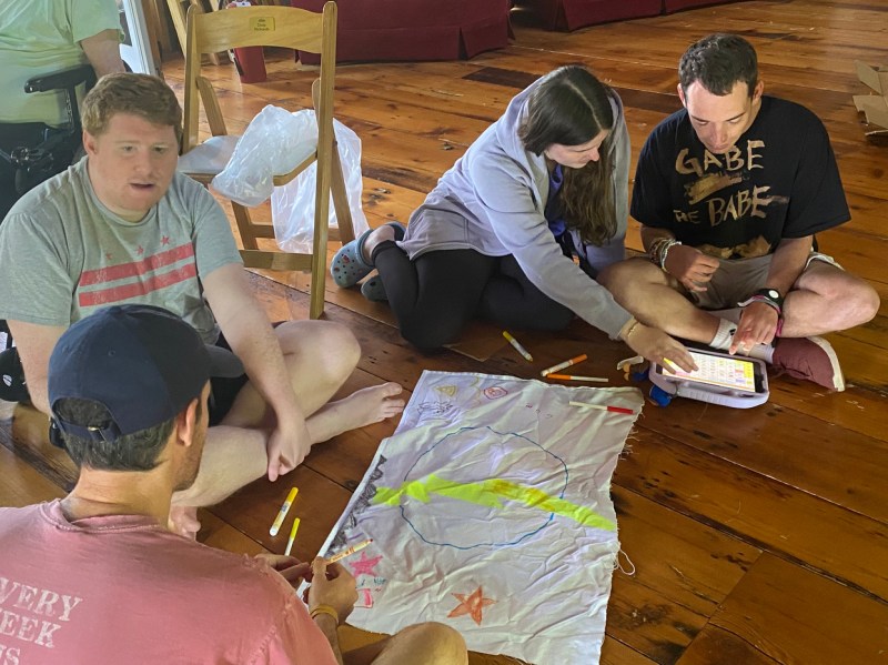 A group of four people sits on a wooden plank four surrounding a rectangular piece of white cloth with drawings of various shapes on it. Two of the people are using an iPad. 