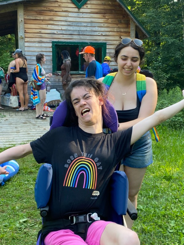 A person sitting in a wheelchair with their arms raised on either side of them. They are wearing a black t-shirt with a rainbow on it that says 'BORN THIS GAY' on it in all capital letters. A person is pushing their chair wearing a black tank top, jean shorts, and a rainbow backpack. 