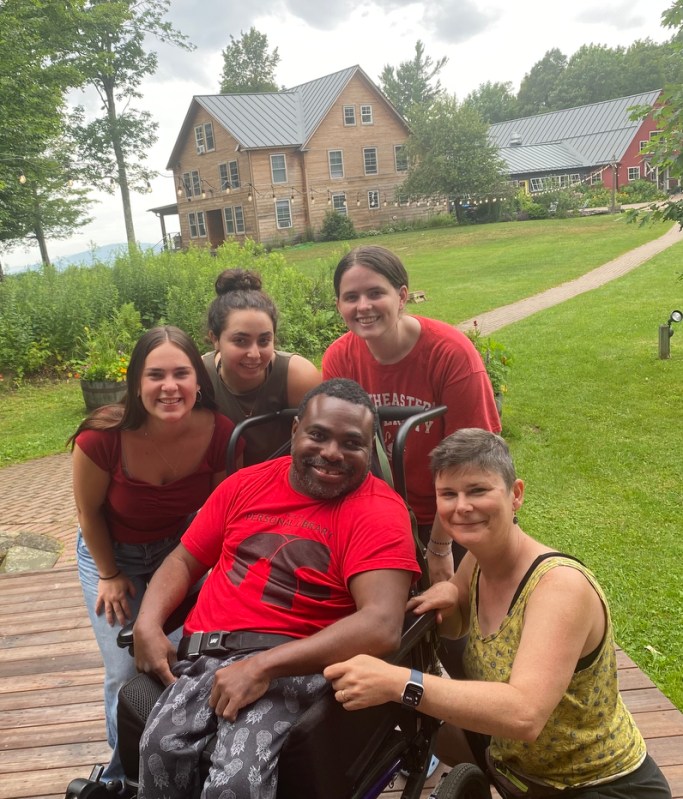 Four people stand and crouch around a fifth person sitting in a wheelchair. They are all smiling for the photo. In the background is a house and string lights strung between trees.