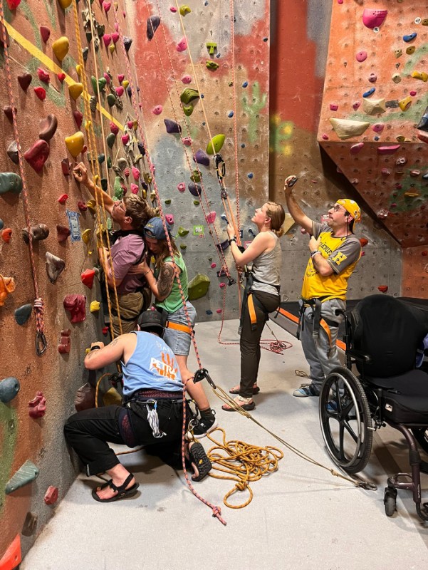 A group of people helps another person rock climb on a wall in an indoor facility. 