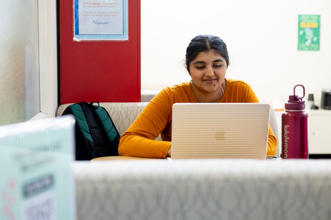 A female Northeastern student with dark hair shown in an orange sweater at work on a laptop.