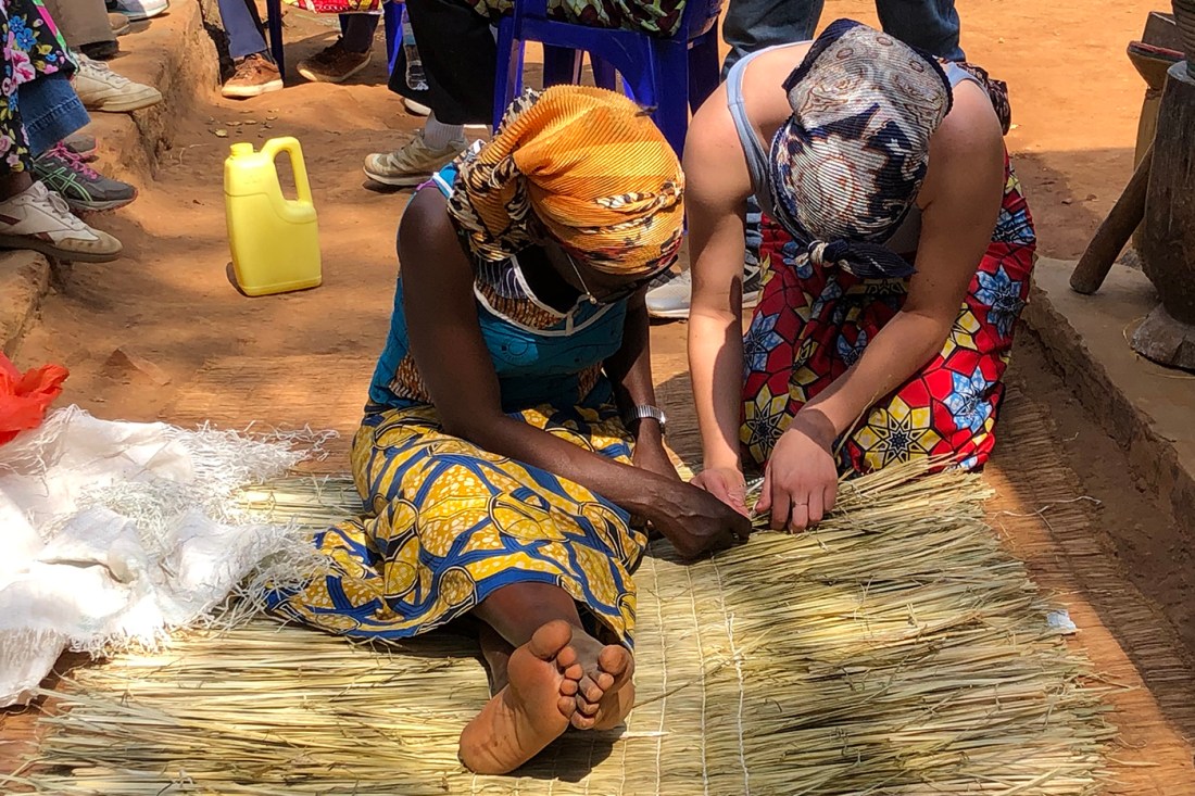Two Black women dressed in traditional garb work together to construct something made of straw.