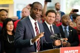 Dr. Joseph Ladapo gesturing while standing in front of a microphone at a podium speaking. He is wearing a suit and tie and standing in front of a crowd of people, including Gov. Ron DeSantis.