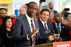 Dr. Joseph Ladapo gesturing while standing in front of a microphone at a podium speaking. He is wearing a suit and tie and standing in front of a crowd of people, including Gov. Ron DeSantis.