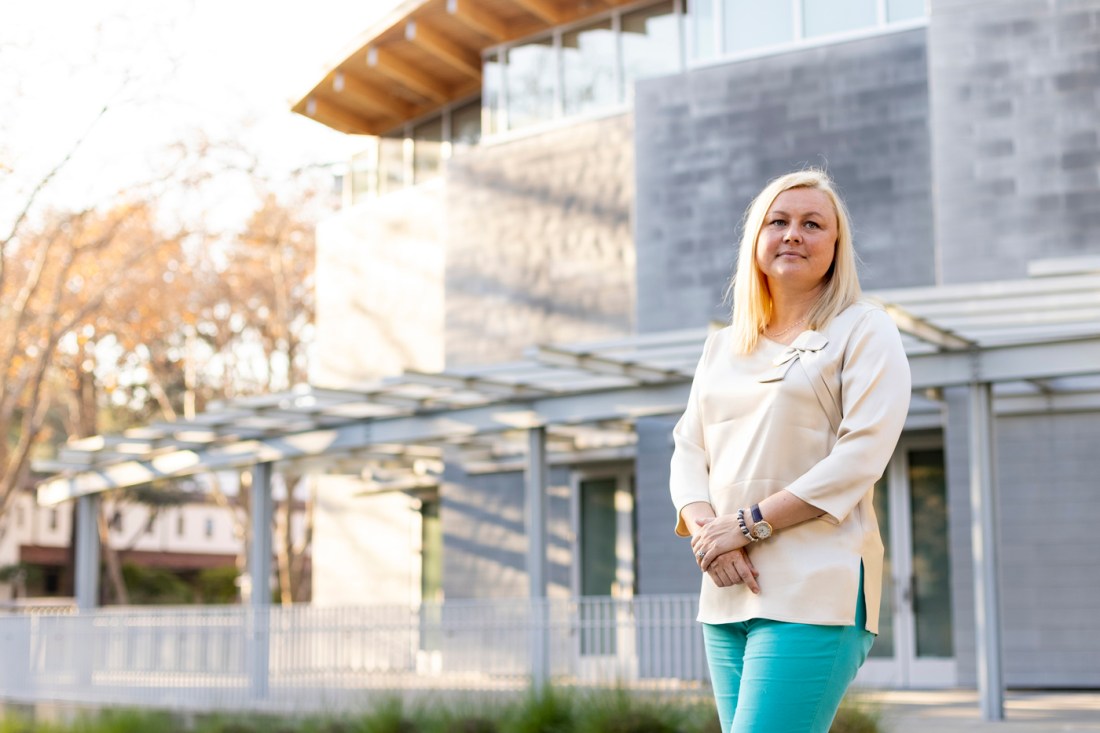 Kate Karniouchina, who is wearing gray-white blouse and teal pants, poses outside of a gray campus building.