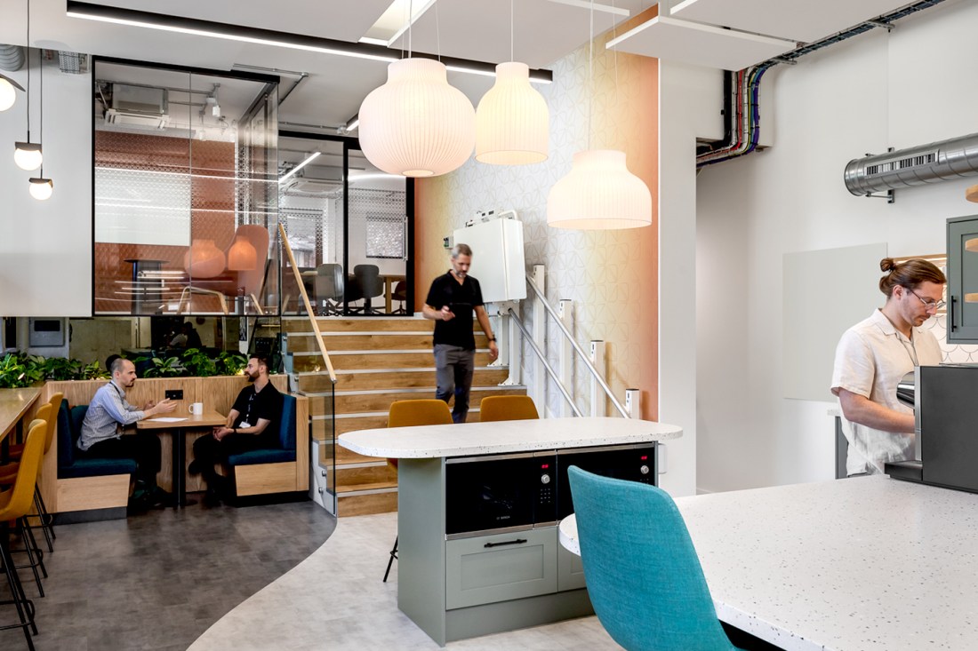 Modern office interior at Marble Quay showing open workspace with wooden tiered seating, white pendant lights, communal kitchen area, exposed ductwork, and staff collaborating in contemporary setting