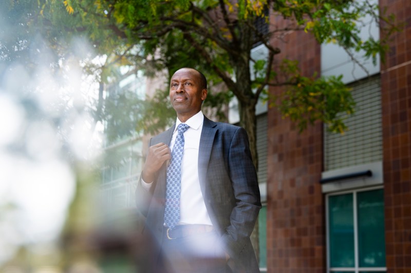 Richard Wamai, a Black professor, standing in front of a tree and brick building wearing a blue suit.