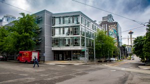A shiny metal and glass building on the Northeastern Seattle campus. There is a red truck and green trees in front, and the Space Needle in the background.