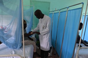 A Black male doctor visits with a Black female patient between blue hospital curtains.