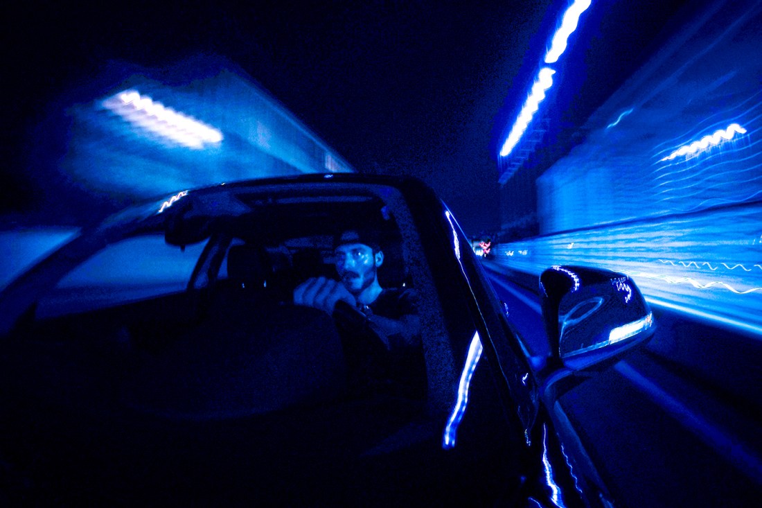 A dark, blurry image of a young bearded man speeding in his vehicle at night.