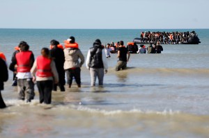 A shot of migrants wading through waters in life vests in the middle of the afternoon.