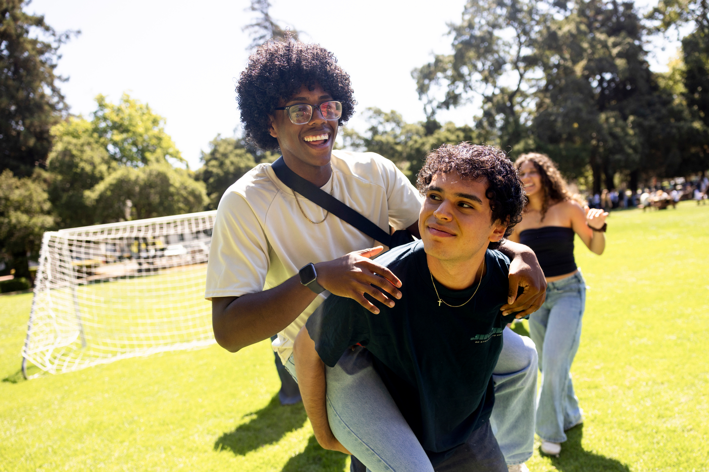 A smiling student on the back of another student stand on a sunny lawn with trees and other people visible in the background.