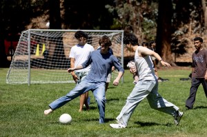 Students compete for a soccer ball on a sunny grass field at Northeastern's Oakland campus, with a goal net visible in the background.