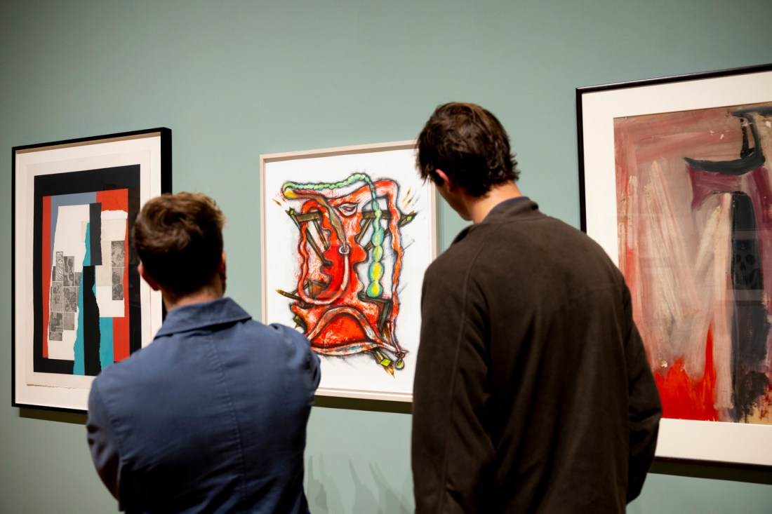 Two museum visitors viewed from behind as they observe framed abstract art pieces hanging on a gallery wall. 