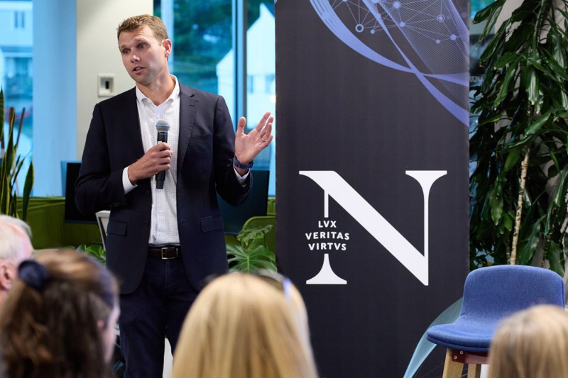 A speaker wearing a button down shirt and blazer holding a microphone and speaking at an event at the Roux INstitute.
