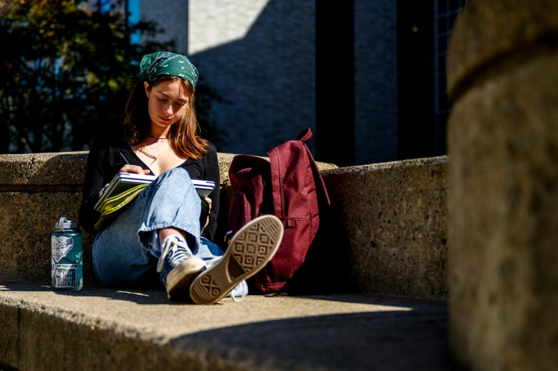 A student does work while sitting on a stone bench on the Boston campus.