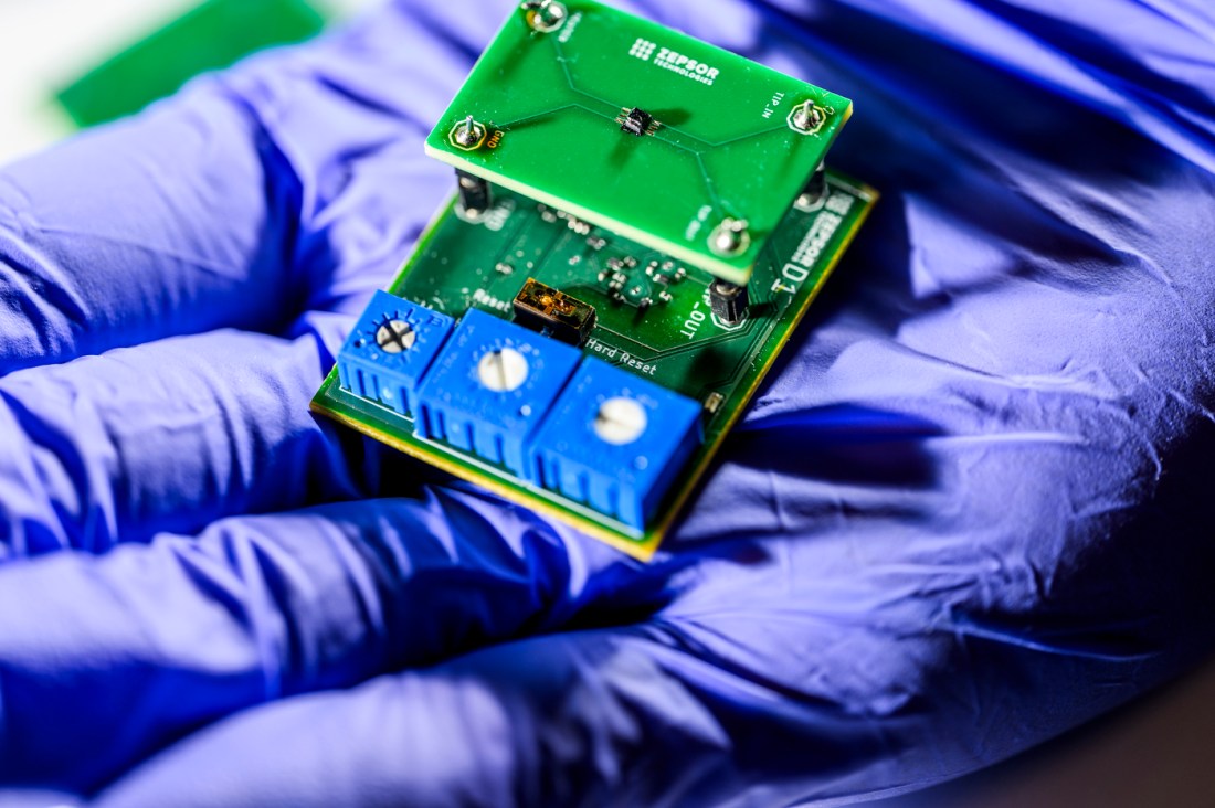A green circuit board is held in a researcher’s gloved hand, shown against a blurred lab background.