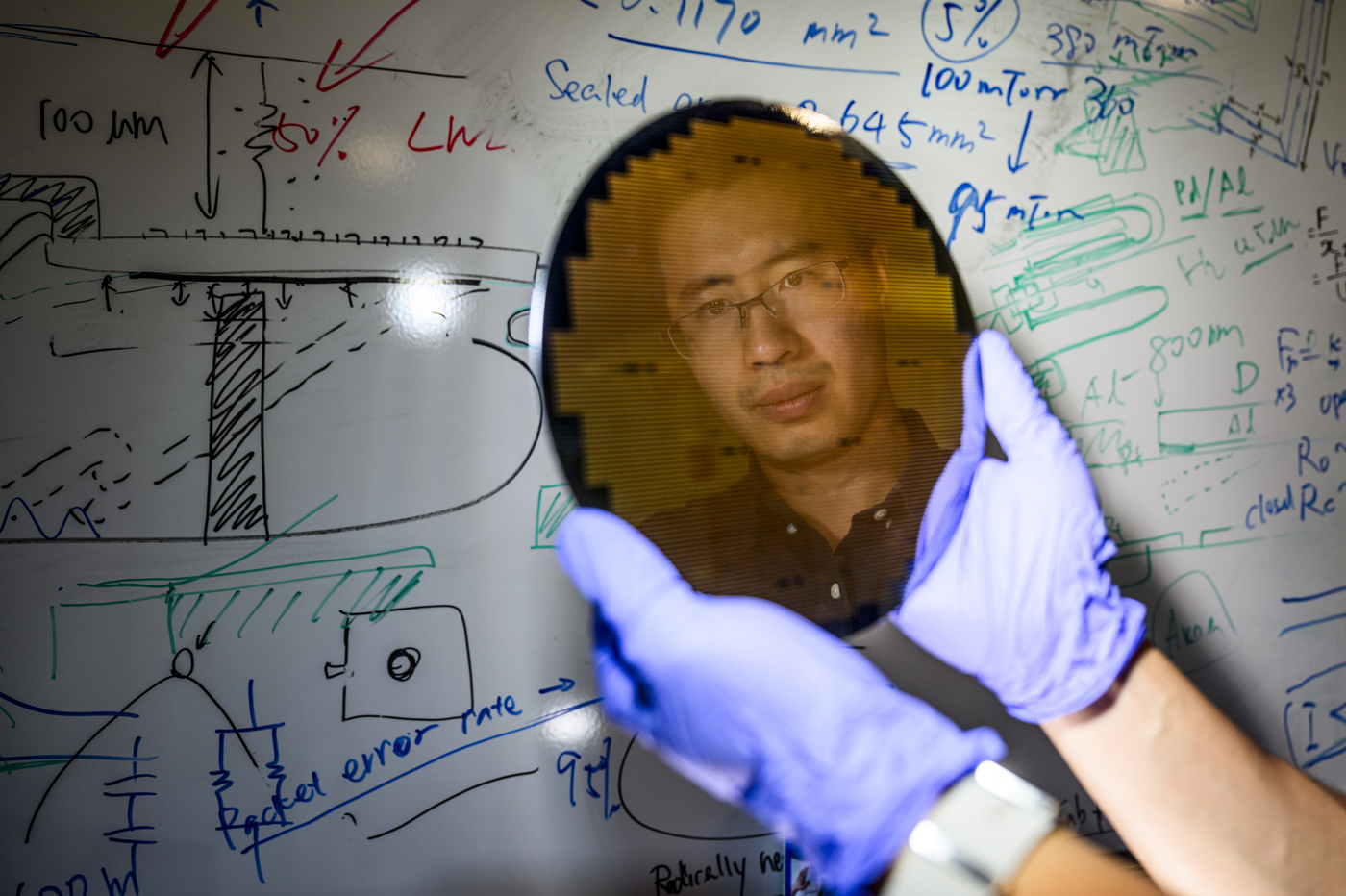 A professor holds a mirror, which shows his reflection, in front of a whiteboard covered in equations.