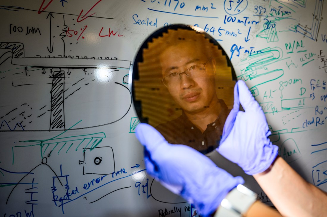 A researcher wearing blue gloves holds up a circular silicon wafer reflecting his face, standing in front of a whiteboard filled with technical drawings and handwritten equations.