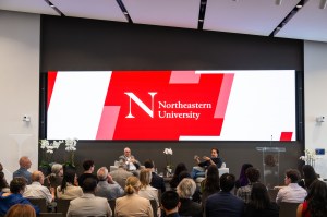 Northeastern University President Joseph E. Aoun sits next to Hemant Taneja on a platform during a fireside chat, with a screen depicting the Northeastern University logo raised above them.