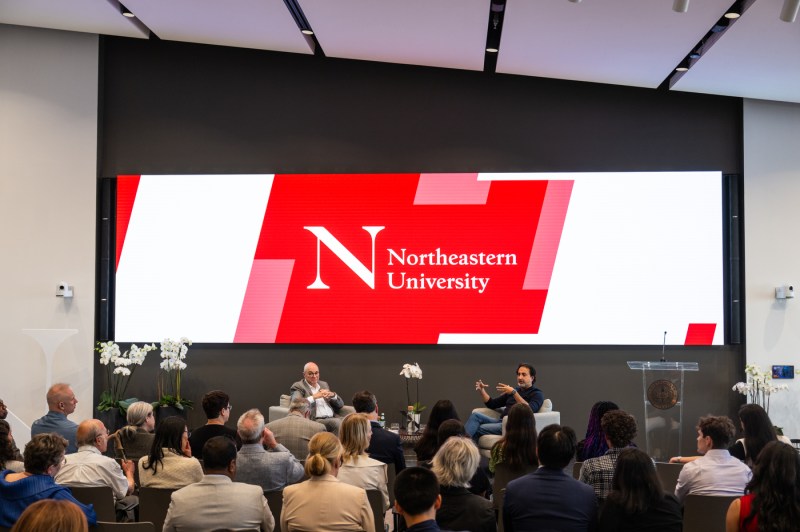 President Aoun and Hemant Taneja sit on chairs on a stage in front of a screen that reads, "Northeastern University," while speaking to a crowd in the audience.