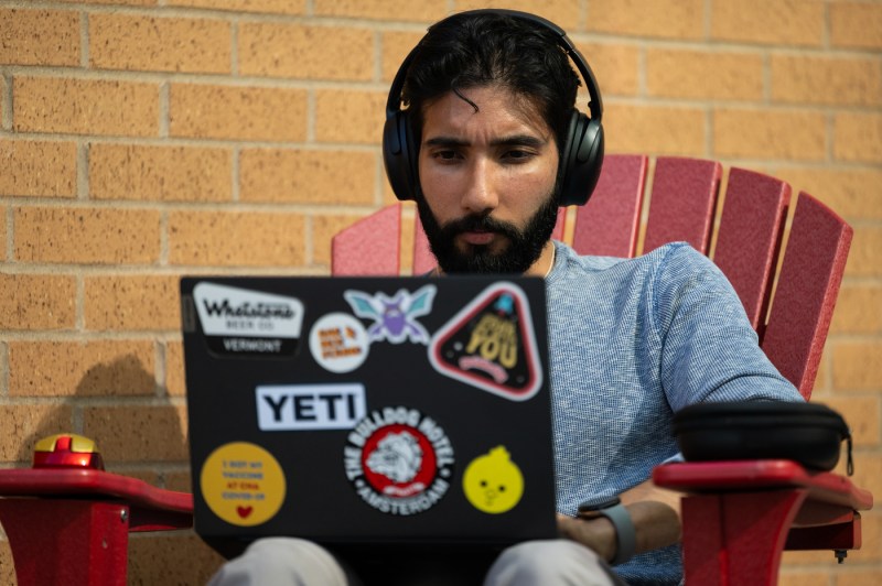 A professor, who is wearing headphones and working on a laptop, sits in a chair outside.