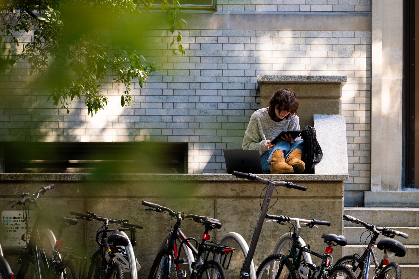 A person sits on a stone wall with a laptop and iPad.