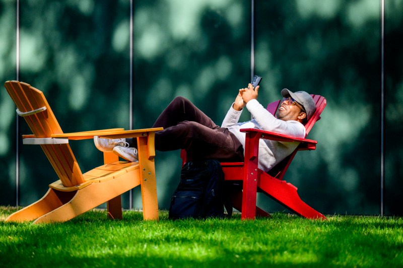 A person sits on an Adirondack chair while looking at their phone and resting their legs on another chair.