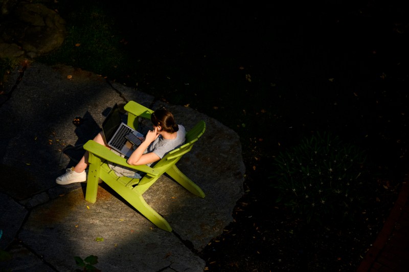 A person sits on a green Adirondack chairs while working on a laptop. 
