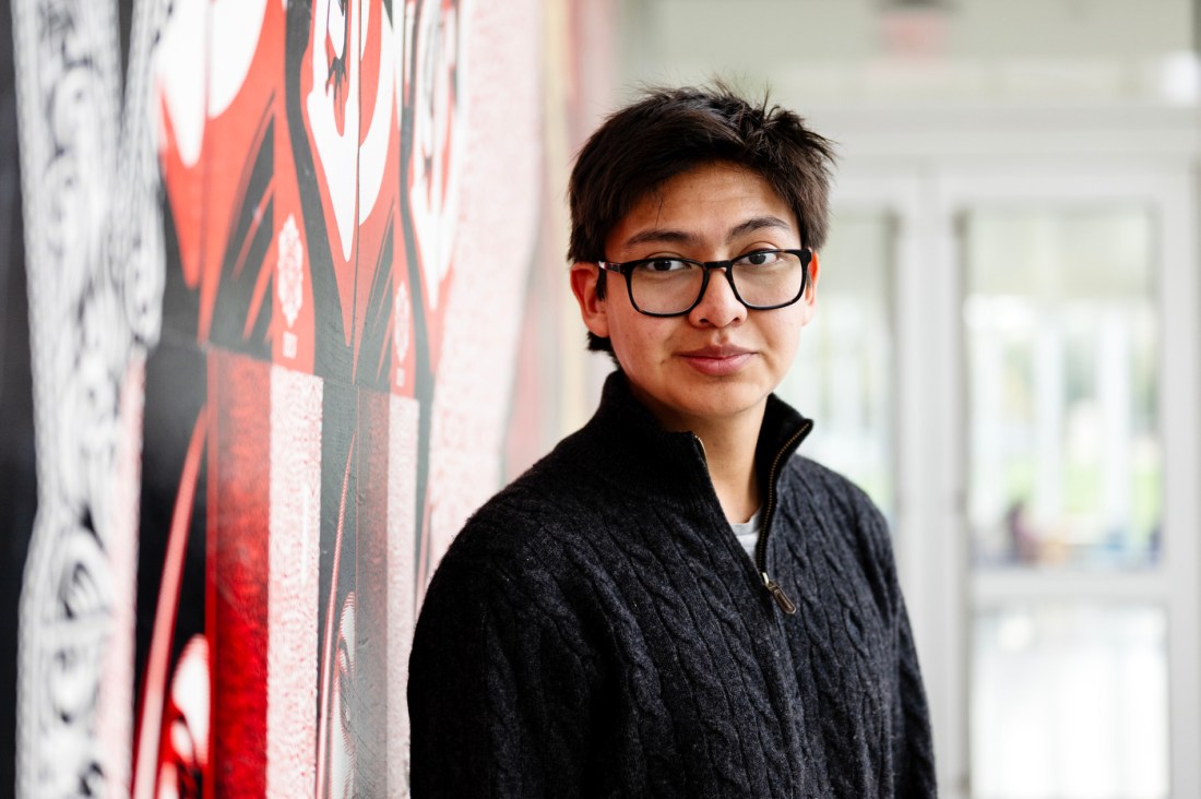 The NATIVE Club Vice President Tamarron Austin, seen in a black sweater with glasses, smiles as he stands just in front of a red, black and white patterned wall. 