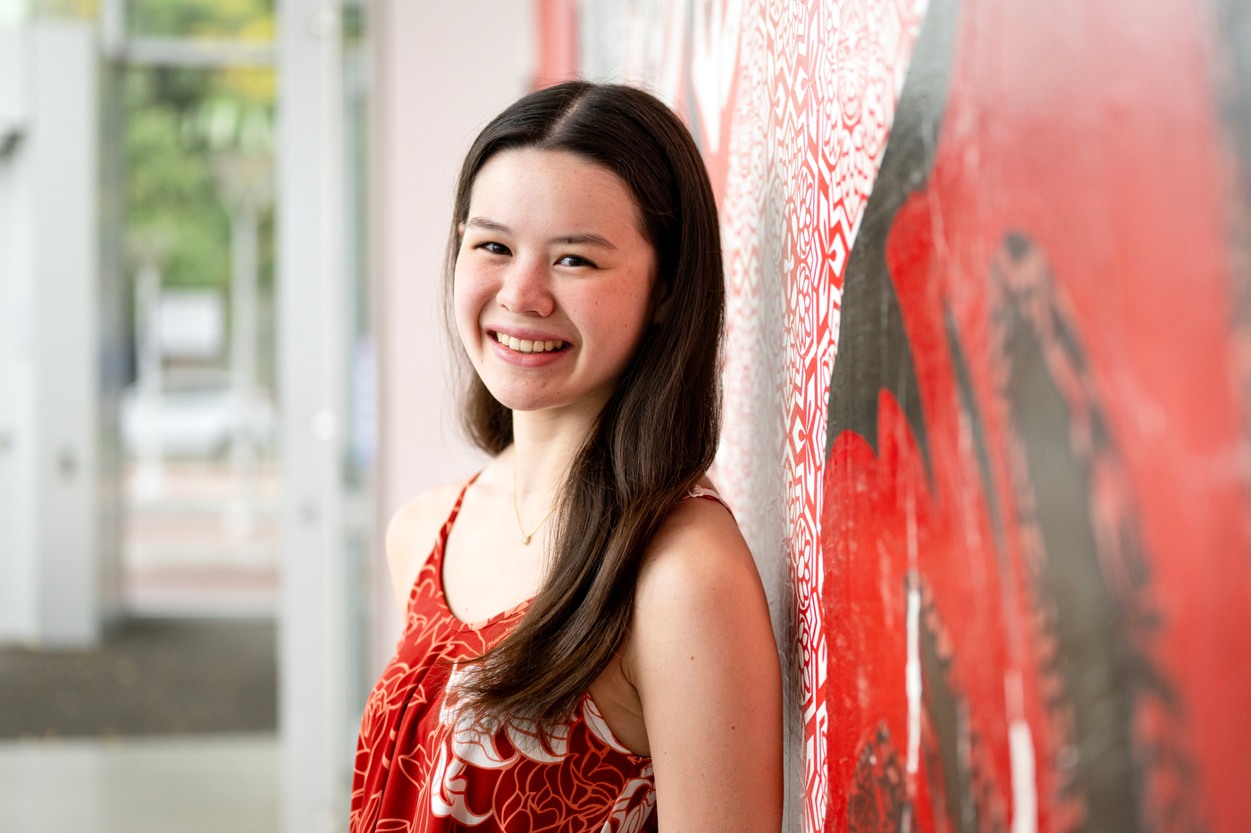 NATIVE student group President Avery Nakata, seen in a red dress and long brown hair, leans against a red and black patterned wall and smiles.