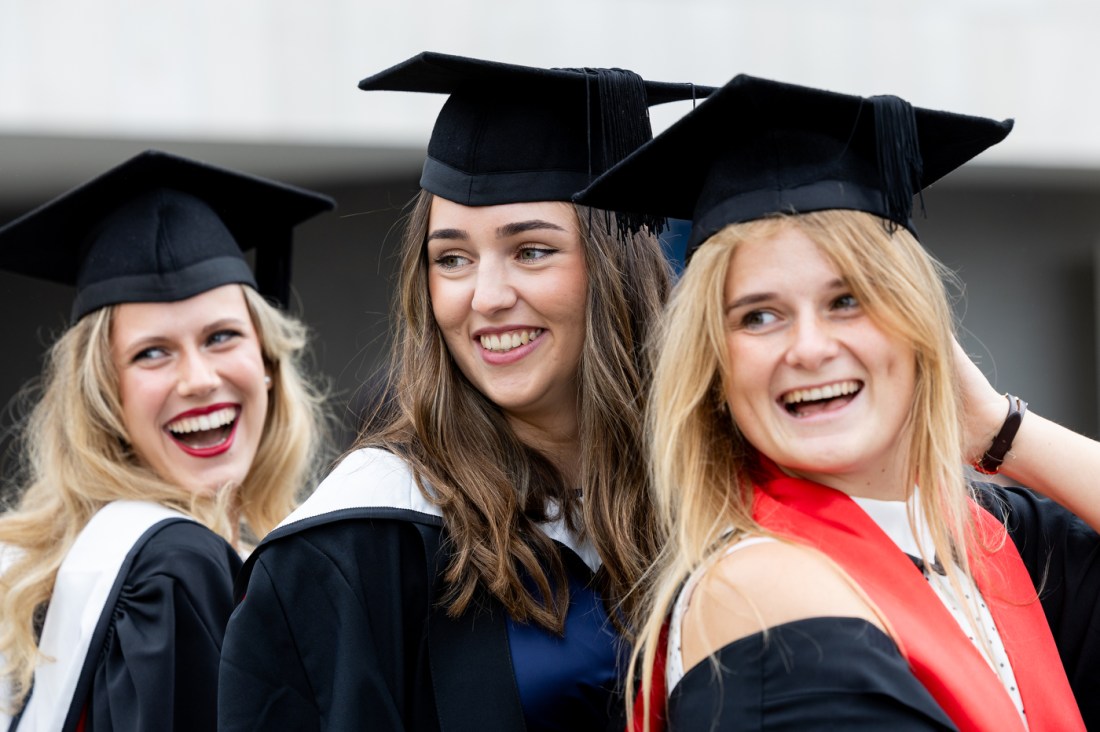 Three graduates in caps and gowns smile and pose together outside Queen Elizabeth Hall during Northeastern University’s London graduation ceremony.