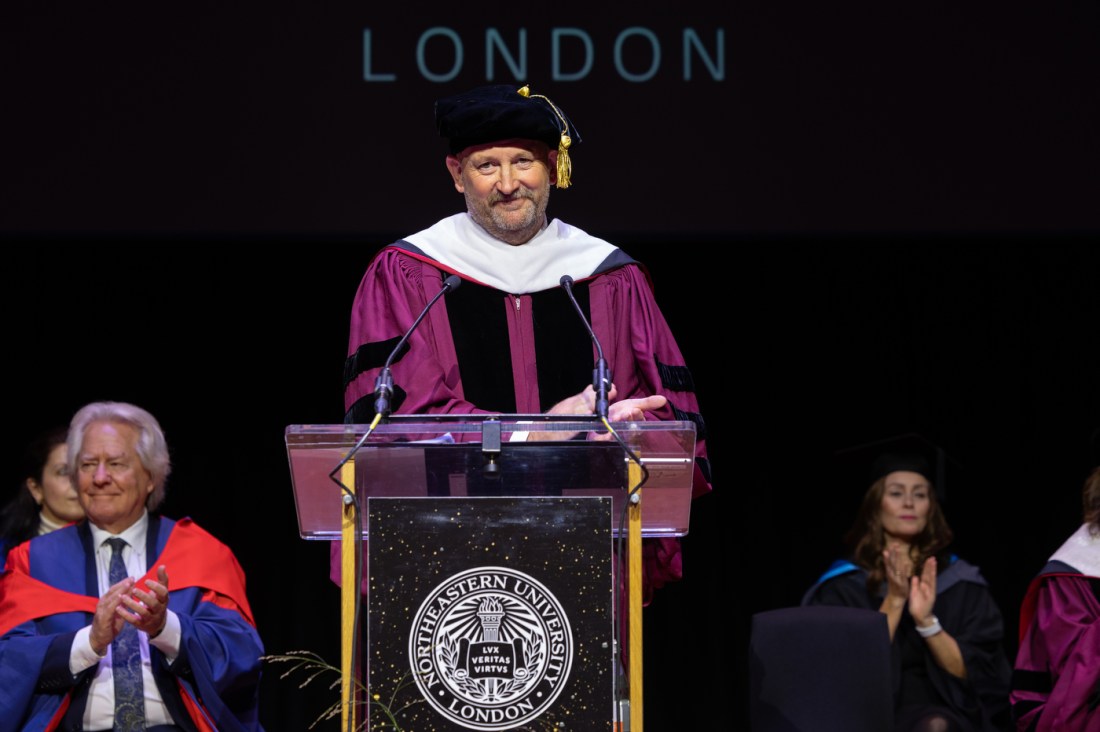 A university official in ceremonial academic robes speaks at a podium during Northeastern University’s London graduation ceremony at Queen Elizabeth Hall.