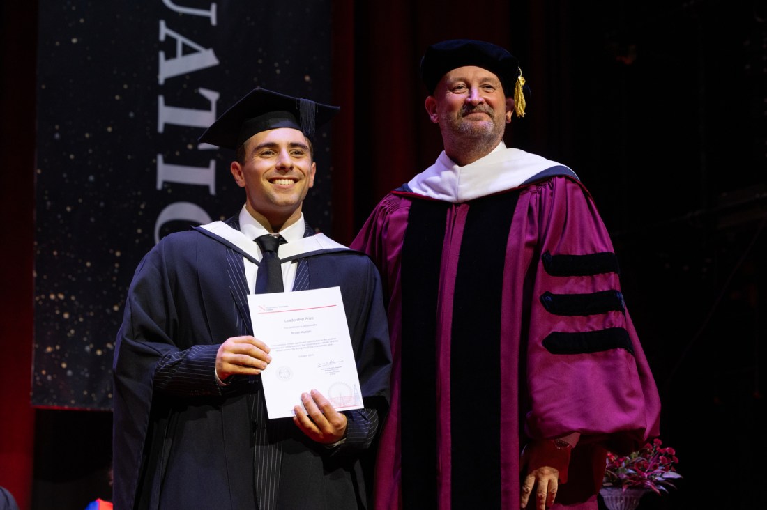 A male student in cap and gown on stage with a male university official to his left, smiling for a photo.