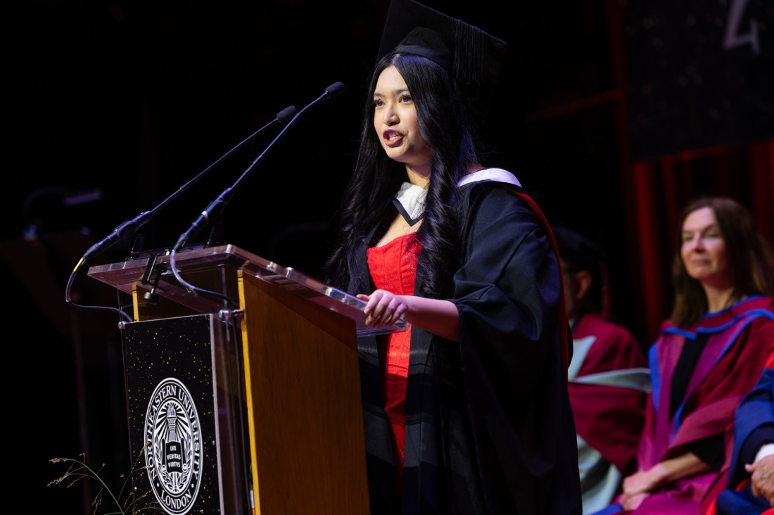 A graduate in a red and black gown speaks at the podium during Northeastern University’s London graduation ceremony.