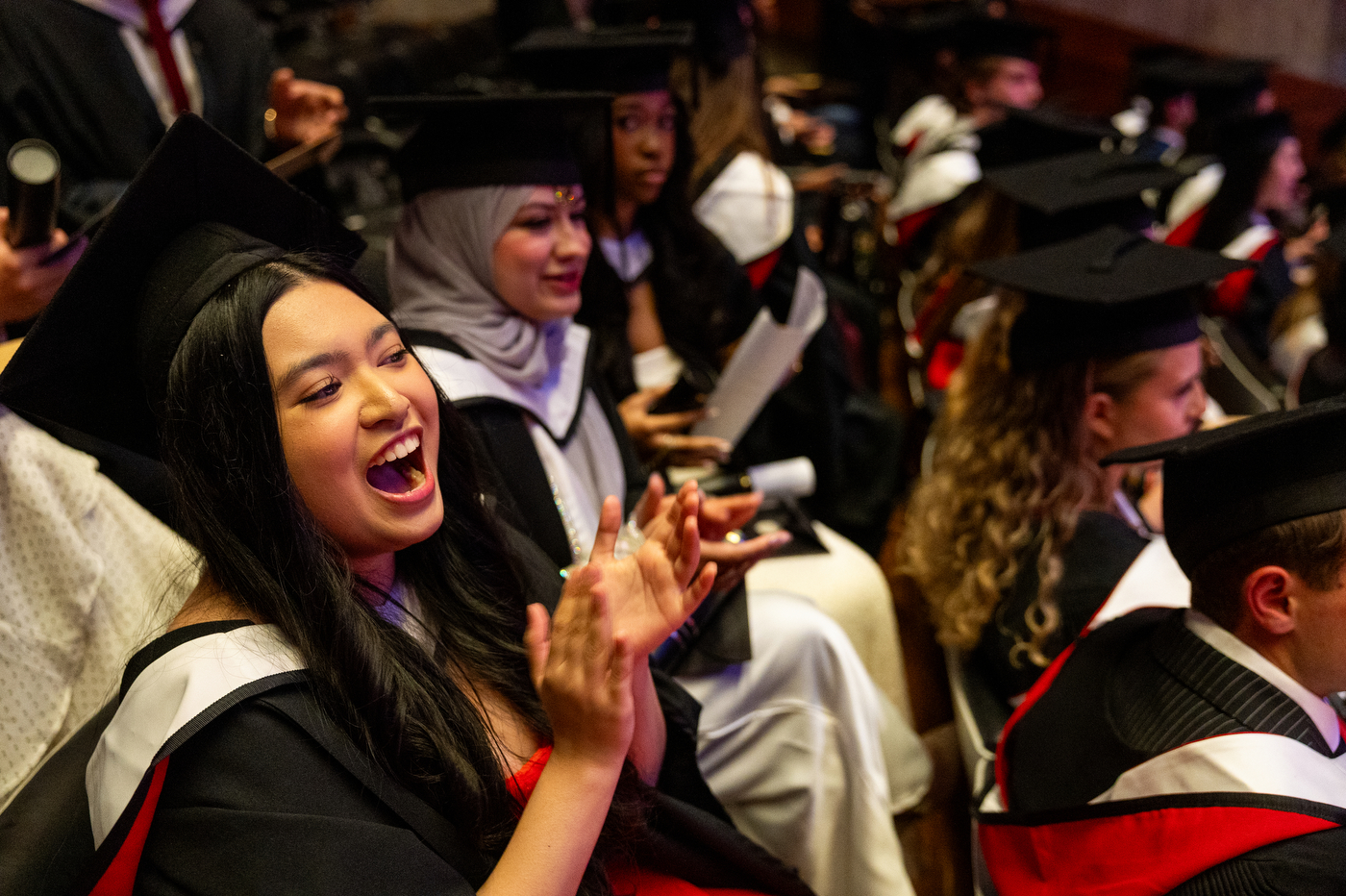A row of graduates in caps and gowns clapping and cheering during the Northeastern London graduation ceremony.