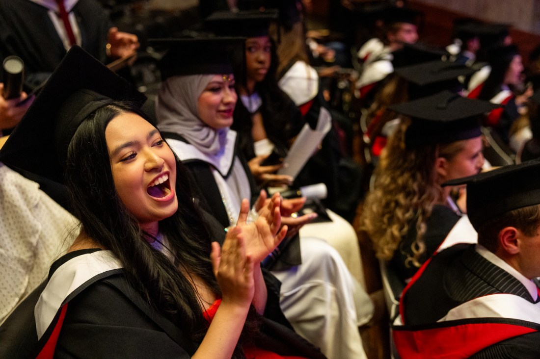 A row of graduates in caps and gowns clapping and cheering during the Northeastern London graduation ceremony.